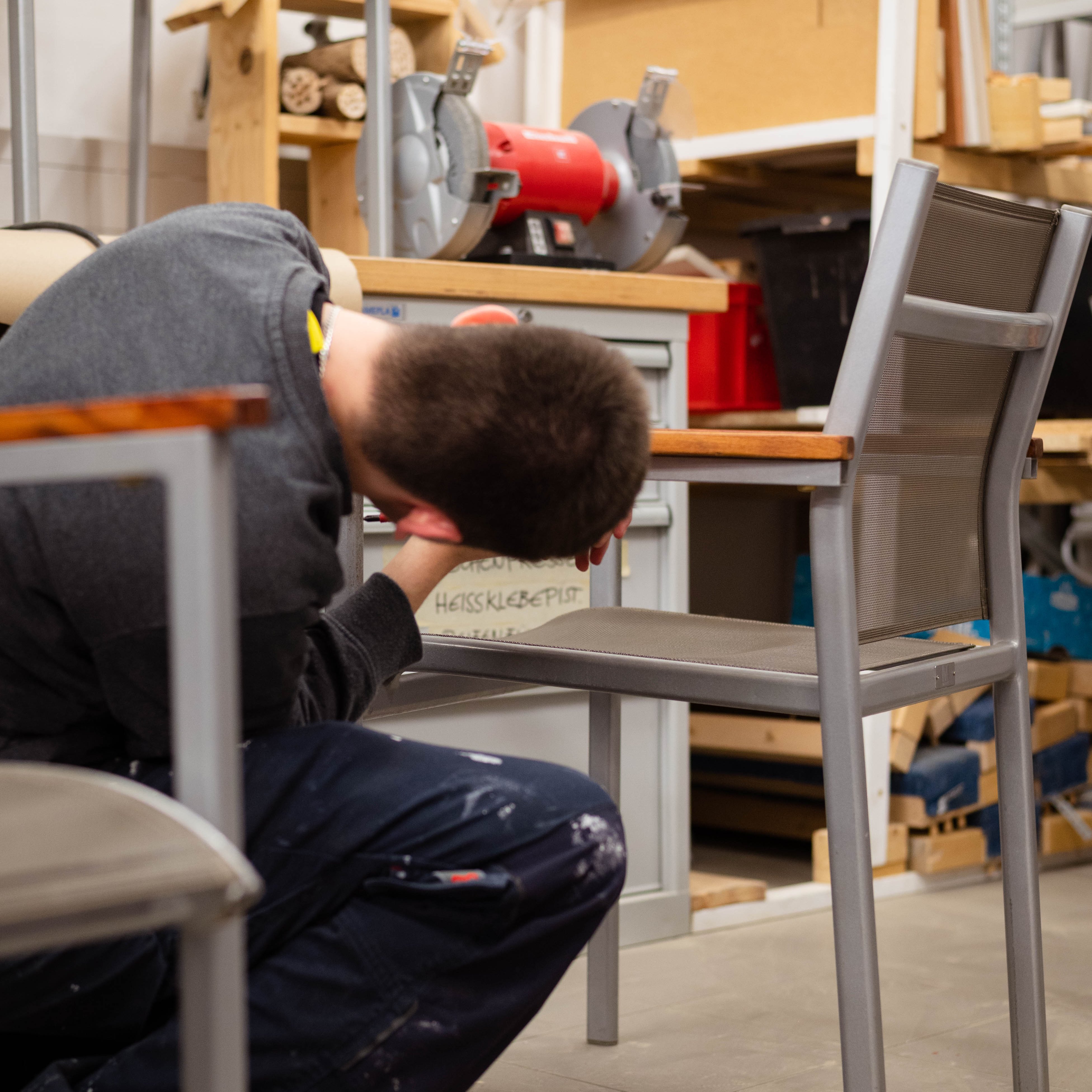Junger Mann arbeitet kniend in einer Werkstatt an der Montage eines Stuhls; im Hintergrund stehen Werkbank, Werkzeug und Maschinen.