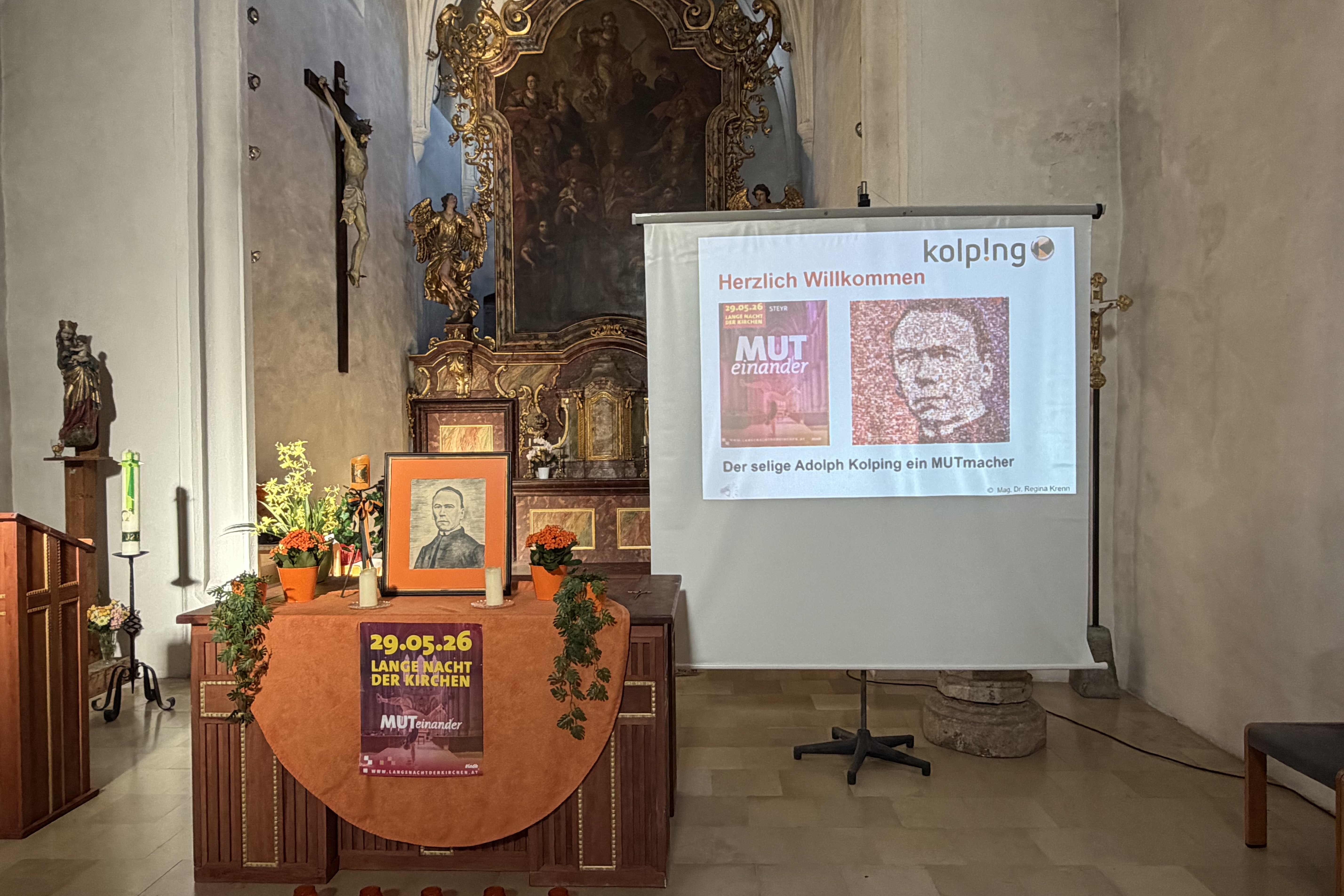 Altarraum einer historischen Kapelle mit Kolping-Präsentation: geschmückter Tisch mit Porträt von Adolph Kolping, Kerzen und Plakat „Lange Nacht der Kirchen“, daneben eine Leinwand mit Begrüßungsfolie zum Thema „MUTeinander“